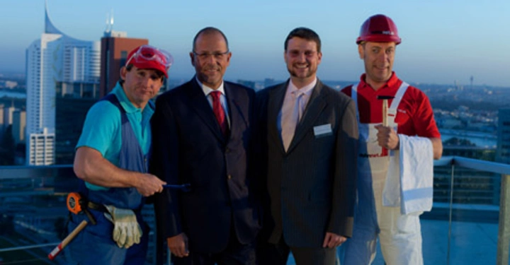 Galerie-Bild 4: Vier Männer stehen auf einer Dachterrasse vor einer Stadtlandschaft Der linke trägt Bauarbeiterkleidung und hält Werkzeug Zwei in der Mitte tragen Anzüge Der rechte trägt Bauarbeiterkleidung und einen Helm