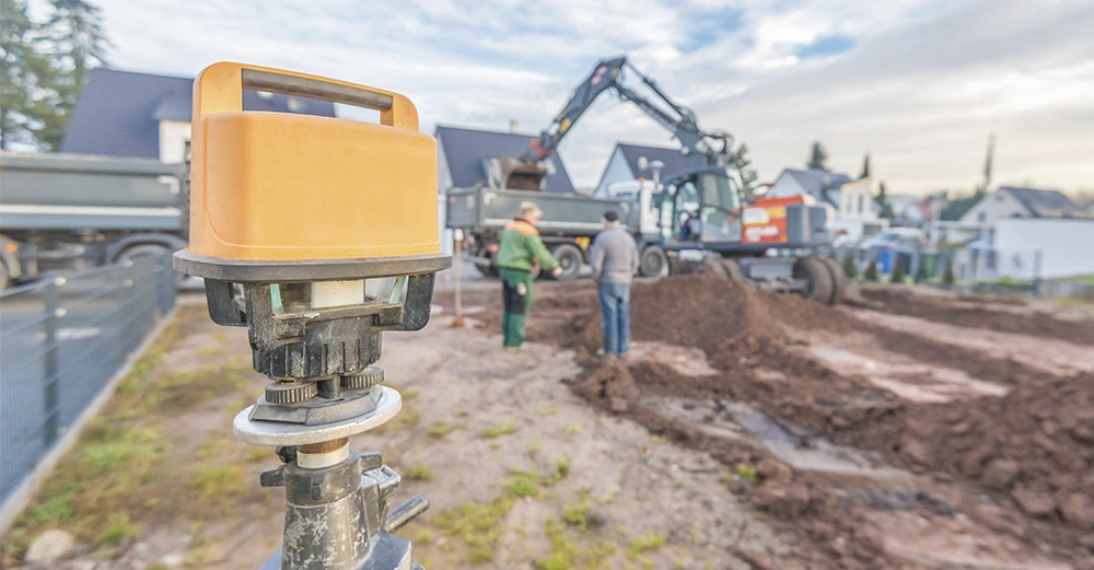 privates Grundstück mit Bagger im Hintergrund, der Erde in einen LKW-Hänger lädt, zwei Männer, die sich unterhalten, im Vordergrund ein Vermessungsinstrument