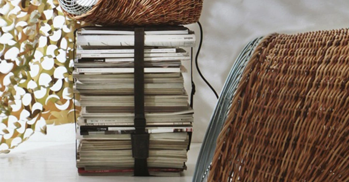 Rattan-Ventilator aktiviert auf gestapelten Zeitschriften in einem hellen Raum mit einem Blattmuster-Vorhang im Hintergrund. Ein weiterer identischer Ventilator liegt auf dem Boden.