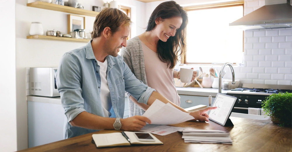 jüngeres Paar steht in Küche und betrachtet Pläne, Frau scheint schwanger zu sein, es soll ein Haus gebaut werden.