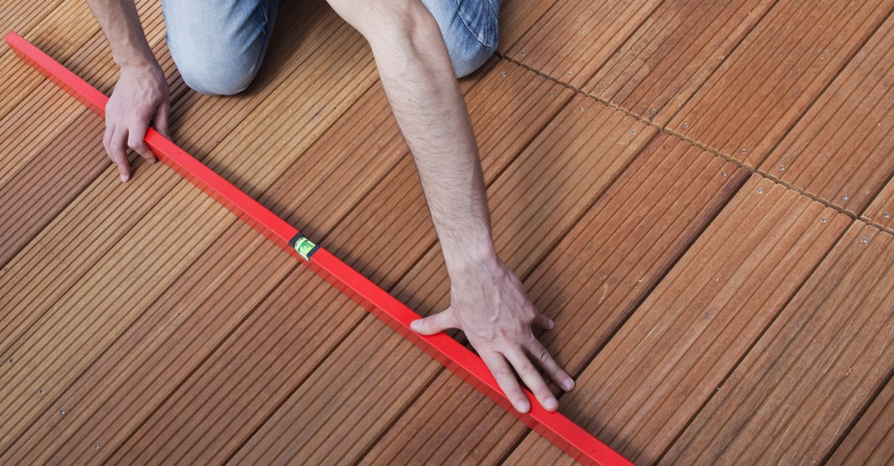 Jemand kniet auf einem Holzdeck und hält eine rote Wasserwaage, um die Ausrichtung der Holzplanken zu überprüfen.