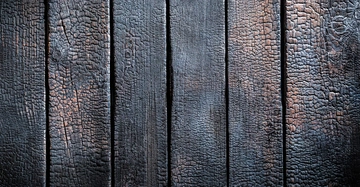 Verkohlte Holzbretter mit einer verbrannten Oberfläche liegen flach nebeneinander. Die Umgebung deutet auf eine strukturelle Anordnung hin und die Textur zeigt deutliche Brandmuster.