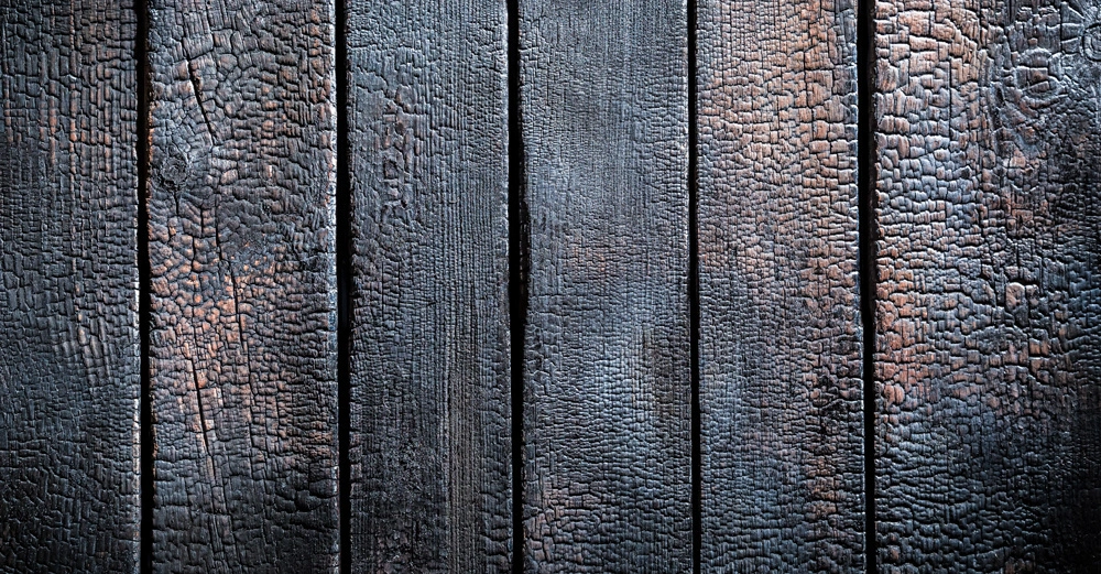 Verkohlte Holzbretter mit einer verbrannten Oberfläche liegen flach nebeneinander. Die Umgebung deutet auf eine strukturelle Anordnung hin und die Textur zeigt deutliche Brandmuster.