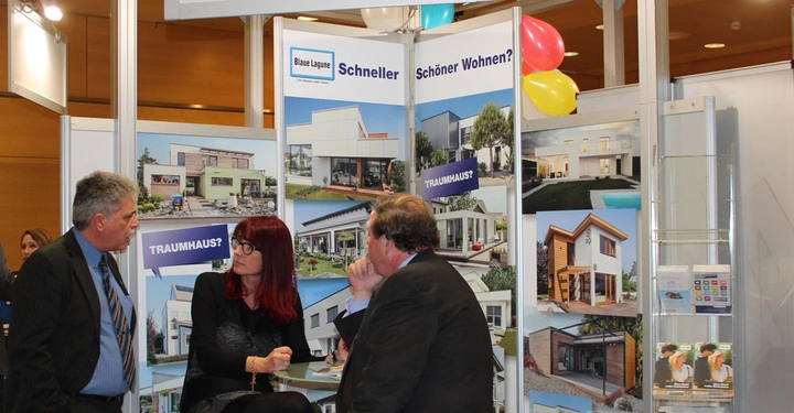 Galerie-Bild 5: Drei Personen unterhalten sich vor einer Ausstellungswand mit Architektur-Postern in einem Messe-Stand Blaue Lagune im Hintergrund Ballons und helle Beleuchtung auf einer Messe Veranstaltung. Text: Blaue Lagune Schneller Schöner Wohnen? TRAUMHAUS? FERTIG!