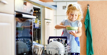 Kleines Kind räumt Geschirrspüler ein sauberer Teller in der Hand in einer Küche mit beigefarbenen Schränken und grünes Handtuch an der Wand hängend.