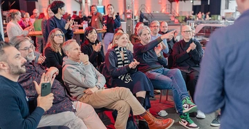 Publikum wirkt interessiert an Vortrag, ein Man zeigt mit dem Arm auf etwas und beugt sich zu seiner Sitznachbarin. Menschen nicht im Businesslook sondern leger gekleidet.