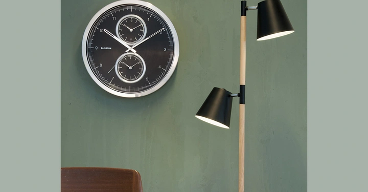 Galerie-Bild 8: Eine runde Uhr mit zwei zusätzlichen Zifferblättern zeigt die Zeit an einer grünen Wand an. Daneben steht eine Stehlampe mit zwei schwarzen Lampenschirmen. Darunter ein Tisch mit Notizbuch und Stift. Text: KARLSSON.