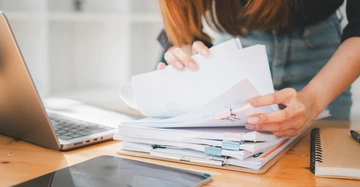 Frau mit langen braunen Haaren sucht in einem Papierstapel nach einem Dokument