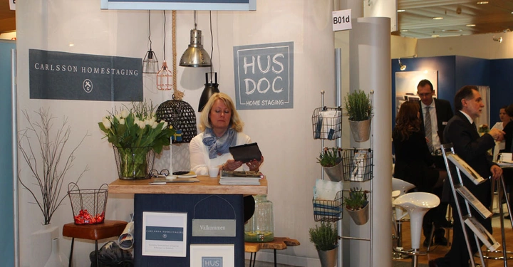 Galerie-Bild 14: Frau sitzt an einem Stand und betrachtet ein Tablet Veranstaltungsumgebung An den Wänden stehen Schilder mit Text Scandianvian HOME STAGING CARLSSON HOMESTAGING HUS DOC HOME STAGING