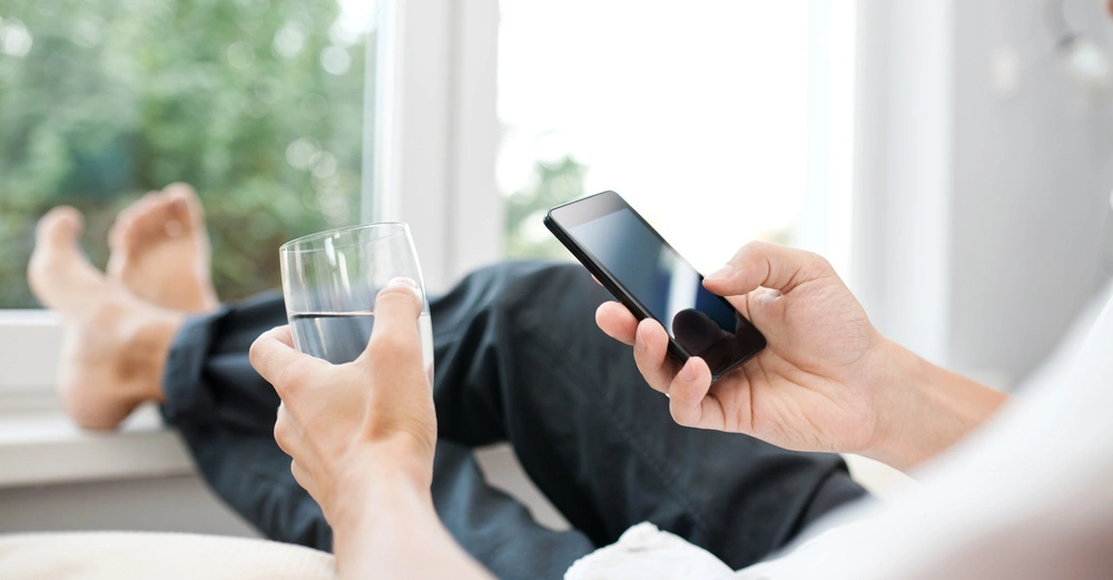 Person hält ein Smartphone in der rechten Hand und ein Wasserglas in der linken Hand während des Entspannens auf einem Sofa bei natürlichem Licht neben einem Fenster.