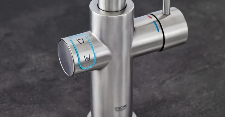 Edelstahl-Wasserhahn zwei Griffe einer mit Symbol für Becher und Wasserglas und der andere für warmes und kaltes Wasser in einer Küche auf einer dunklen Steinoberfläche. Text Grohe.