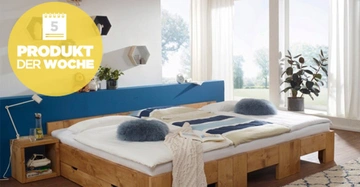 Holzbett mit weißer Bettwäsche und blauen Kissen, steht in einem hellen Schlafzimmer mit großen Fenstern, einer blauen Rückwand und Pflanzen. Text im gelben Kreis: "5 PRODUKT DER WOCHE".