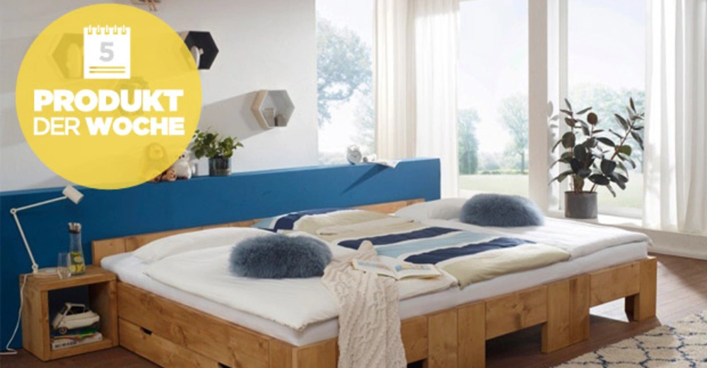 Holzbett mit weißer Bettwäsche und blauen Kissen, steht in einem hellen Schlafzimmer mit großen Fenstern, einer blauen Rückwand und Pflanzen. Text im gelben Kreis: "5 PRODUKT DER WOCHE".