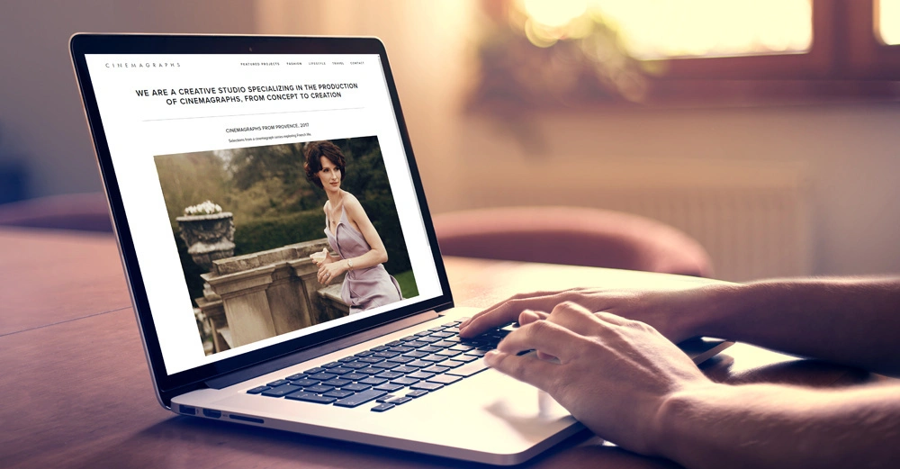 Ein Laptop zeigt eine Webseite über ein Kreativstudio für Cinemagraphs person tippt darauf helle häusliche Umgebung. Text: WE ARE A CREATIVE STUDIO SPECIALIZING IN THE PRODUCTION OF CINEMAGRAPHS FROM CONCEPT TO CREATION CINEMAGRAPHS FROM PROSPEKT 2017.