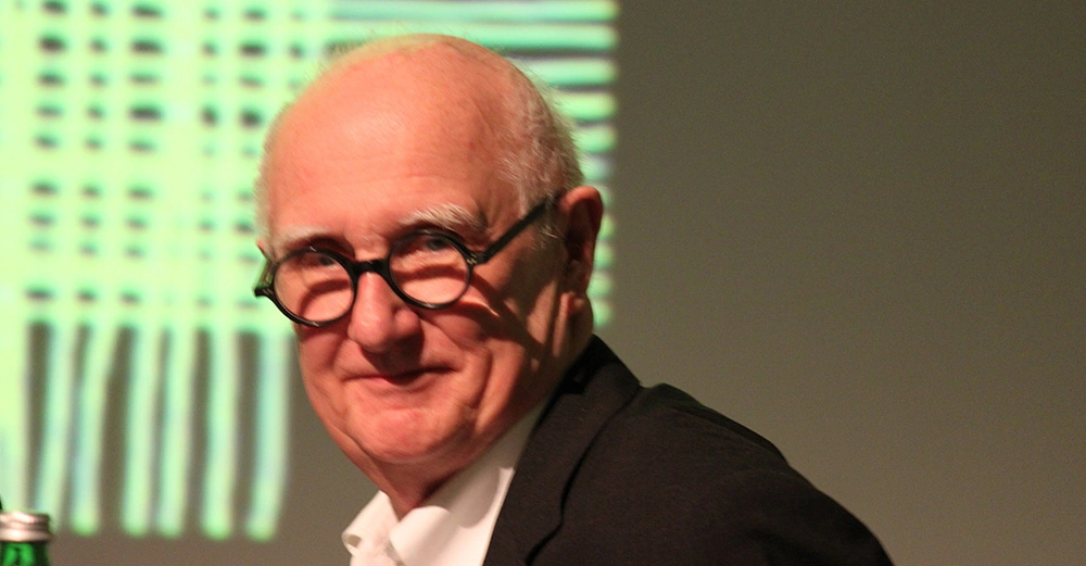 Mann trägt schwarzes Sakko und runde Brille lächelt in die Kamera unscharfer Hintergrund mit vertikalen Linien grüne Flasche im Vordergrund links.