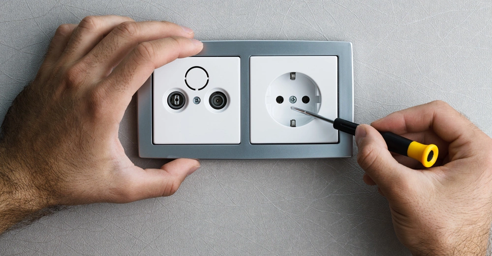Hände montieren eine Steckdose an der Wand, wobei eine Hand sie hält und die andere einen Schraubendreher benutzt. Der Hintergrund besteht aus einer grauen Tapete.