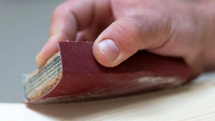 Großaufnahme einer rechten Hand, die mit rotem Schleifblock Holz abschleift. Staub deutlich zu sehen.