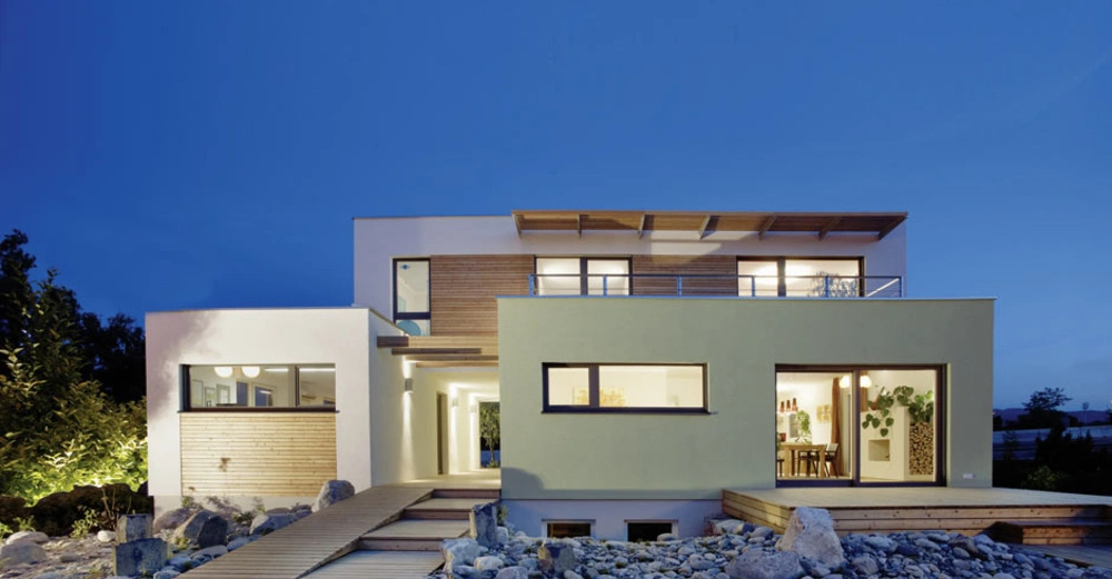 Modernes zweistöckiges Haus beleuchtet in der Dämmerung umgeben von einem steinigen Garten und einem Holzweg mit großen Fenstern die ins Innere blicken lassen.