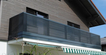 Ein moderner Balkon mit horizontalen Metallstäben über einem weißen Haus mit grüner Markise vor einem mit Holz verkleideten Hauswand im Wohngebiet.