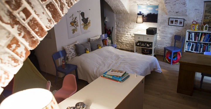 Galerie-Bild 9: Ein großes Bett mit weißen Kissen steht in einem gemütlichen Raum mit weiß gestrichenen Ziegelwänden rundherum. Ein Nachttisch mit Lampe daneben. Ein Bücherregal und bunte Stühle im Raum.