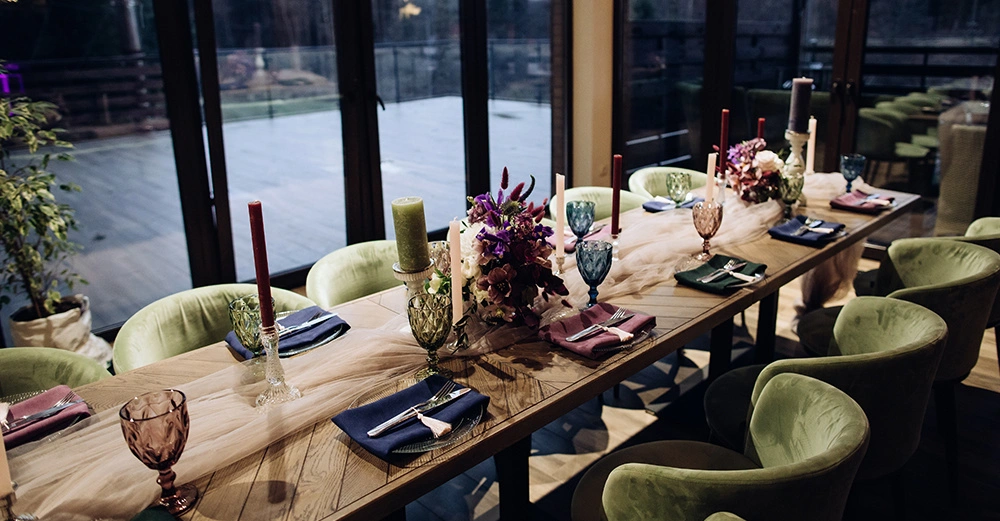 Tisch mit bunten Gläsern und Kerzen Geschirr und Blumenarrangements in einer modernen Umgebung grüne Stühle und große Fenster bieten Blick auf eine Terrasse und Außengegend abends oder bei schwachem Licht