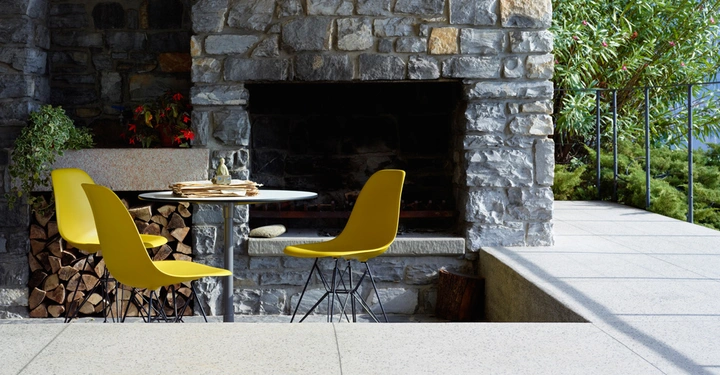 Drei gelbe Stühle und ein runder Tisch mit Zeitschriften stehen vor einem großen Steinkamin. Außenbereich mit Pflanzen und Gehweg.