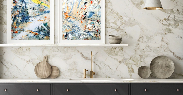 Küchenarbeitsplatte mit hellen Marmorrückwänden zeigt eine goldene Wasserarmatur und dekorative Holzbretter darauf. Darüber hängen zwei abstrakte Gemälde zwischen Regalen mit Keramikschalen.