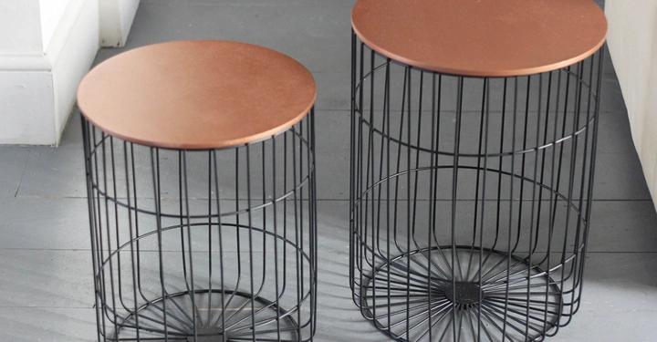 Galerie-Bild 5: Zwei runde Beistelltische mit kupferfarbenen Oberflächen und schwarzen, drahtkorbartigen Gestellen stehen auf einem grauen Holzboden in der Ecke eines Zimmers.