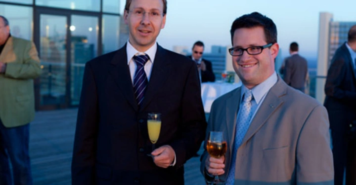 Galerie-Bild 19: Zwei Männer in Anzügen halten Getränke und lächeln auf einer Dachterrasse bei einer formellen Veranstaltung in der Abenddämmerung. Im Hintergrund sind weitere Personen und Gebäude zu sehen.
