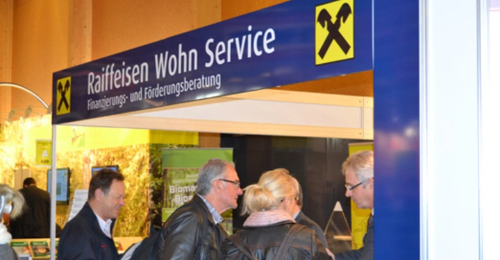 Galerie-Bild 34: Mehrere Personen stehen an einem Informationsstand unter einem Schild mit der Aufschrift Raiffeisen Wohn Service Finanzierungs- und Förderungsberatung in einer hellen, gut besuchten Messeumgebung.