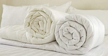 Zwei zusammengerollte weiße Bettdecken liegen nebeneinander auf einem frisch bezogenen Bett mit weißen Kissen im Hintergrund.