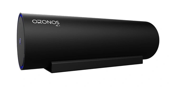 Ein schwarzer zylindrischer Luftreiniger mit der Aufschrift Ozonos AC-1 liegt auf einem schwarzen Ständer in einem isolierten Raum.
