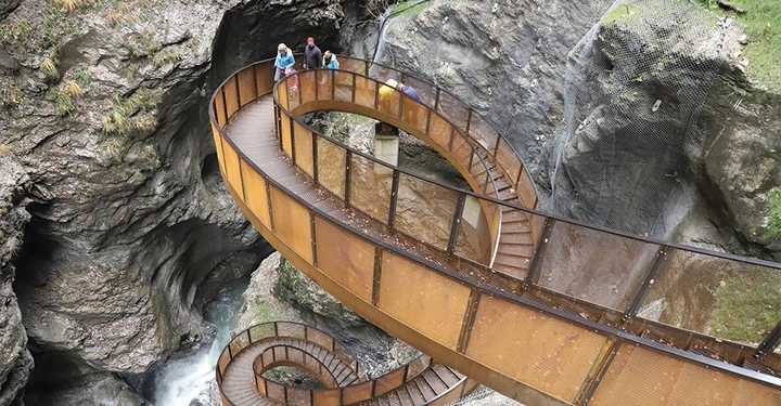 Galerie-Bild 7: Metallische, spiralförmige Fußgängerbrücke schlängelt sich durch steile Felswände; zwei Erwachsene und ein Kind laufen darauf und blicken in die steinige Schlucht mit fließendem Wasser darunter.