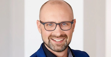 Kahlköpfiger Mann mit Brille und Bart lächelt in einem gut beleuchteten, modernen Innenraum.