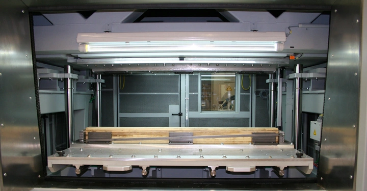 Metallrahmen hält ein Brett horizontal fest umgeben von Maschinenkomponenten in einem industriellen Prüfstandszimmer. Die Umgebung ist mechanisch und funktional erkennbar an Schienen und Steuerungskabeln.