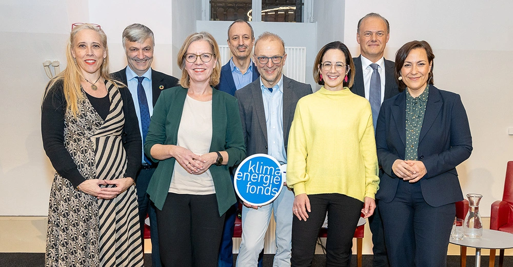 acht Personen (4 Männer, 4 Frauen), darunter Leonore Gewessler stehen auf einer kleinen Bühne, im Hintergrund ein Fenster, ein Mann im Vordergrund hält ein kleines Schild des Klilma- und Energiefonds in die Kamera