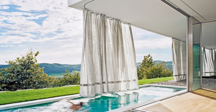 Galerie-Bild 11: Person schwimmt in einem schmalen Pool zwischen modernen Gebäudewänden auf einer Terrasse mit Vorhängen im Vordergrund und bewaldetem Hügel im Hintergrund unter bewölktem Himmel.
