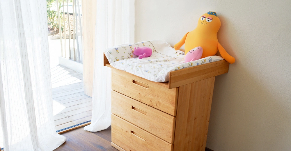 Hölzerne Kommode mit Wickelauflage darauf drei Stofftierfiguren stehend in einem hellen Raum mit weißen Vorhängen und Holzboden die Außenterrasse mit Pflanzen ist durch eine Glastür sichtbar