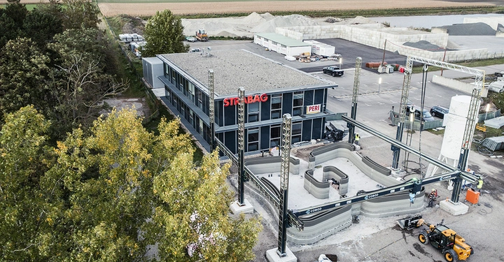 Industriegebäude mit STRABAG und PERI-Logos große 3D-Drucker drucken Betonstrukturen im Außenbereich umgeben von Bäumen und einer Baustellenumgebung mit Fahrzeugen und Materialien im Hintergrund