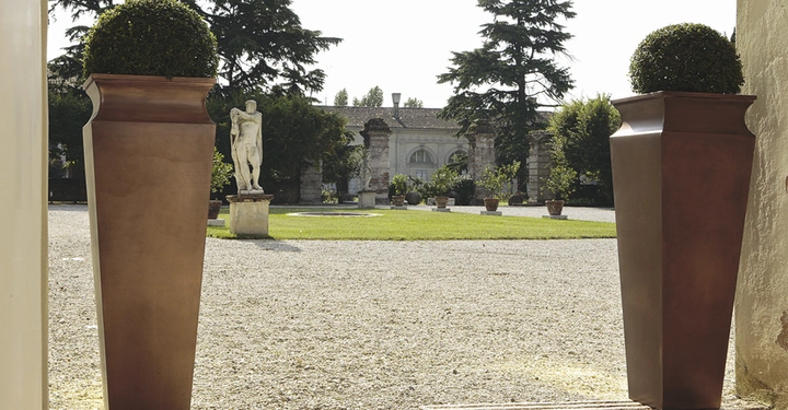 Galerie-Bild 2: Zwei hohe, braune Pflanzgefäße mit kugelförmigen Buchsbäumen stehen auf einem Kiesweg mit einer Rasenfläche und einer Statue im Hintergrund, umgeben von Bäumen und historischen Gebäuden.