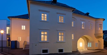 Ein großes weißes Gebäude mit beleuchteten Fenstern steht in einer städtischen Umgebung bei Dämmerung. Eine Straßenlaterne beleuchtet den Gehweg daneben.