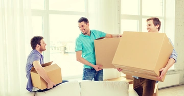Drei Männer tragen Kartons ein Mann sitzt mit einem Karton auf dem Sofa die anderen beiden stehen und lächeln in einem sonnigen hellen Raum mit großen Fenstern im Hintergrund.