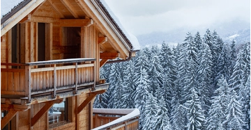 Holzchalet mit Balkon inmitten eines verschneiten Waldes in den Alpen, umgeben von schneebedeckten Tannen und winterlicher Berglandschaft.
