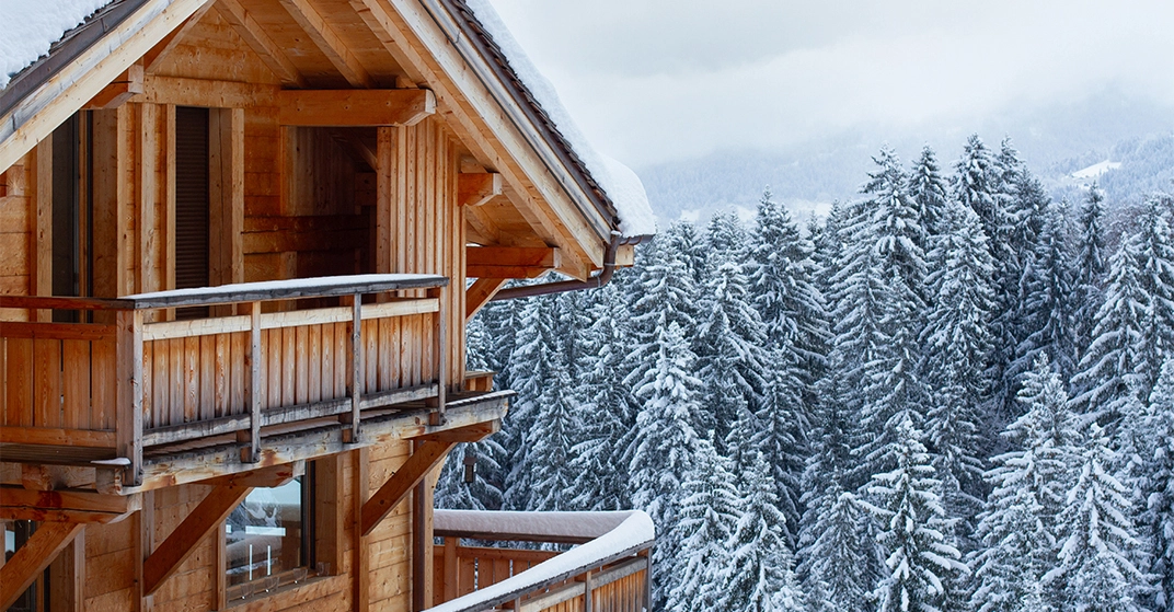 Holzchalet mit Balkon inmitten eines verschneiten Waldes in den Alpen, umgeben von schneebedeckten Tannen und winterlicher Berglandschaft.