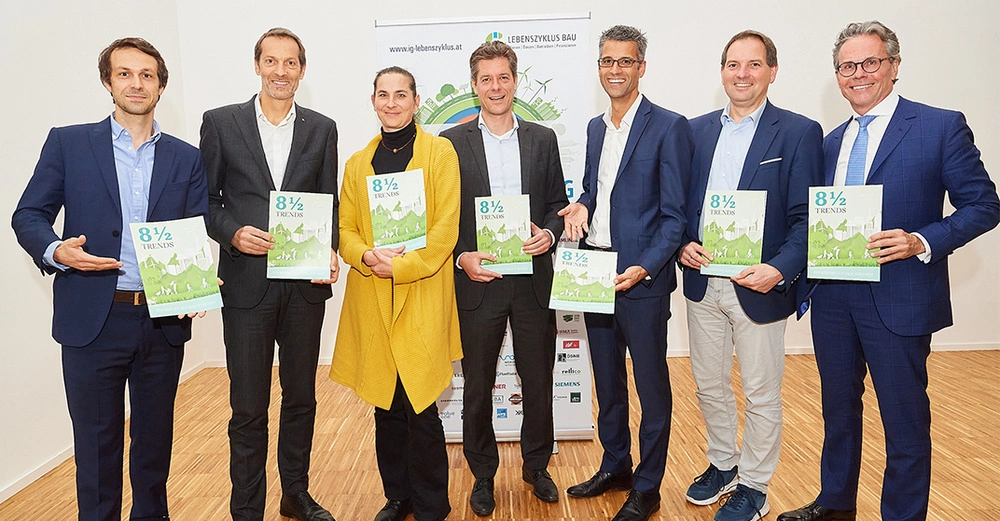 Sieben Personen stehen nebeneinander und halten grüne Broschüren mit dem Titel 8½ Trends in Händen ein Plakat von www.ig-lebenszyklus.at im Hintergrund helle Raumumgebung Holzboden.
