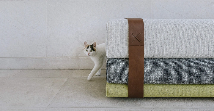 Eine weiße Katze geht neben einem Stapel von Stoffen in verschiedenen Farben und Texturen, die mit einem braunen Lederriemen zusammengehalten werden, in einem minimalistisch gestalteten Raum.