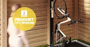 Ein Fahrrad steht vertikal an einem Ständer innerhalb eines Holzschuppens. Ein unscharfer Mensch geht vorbei. Text: 2 Produkt der Woche.