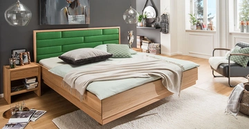 Ein Bett mit grünem Kopfteil steht in einem modernen Schlafzimmer mit Holzboden und grauer Wand; Tagesdecke ist beigefarben, zwei Kissen sind grau und grün gemustert. Rug.