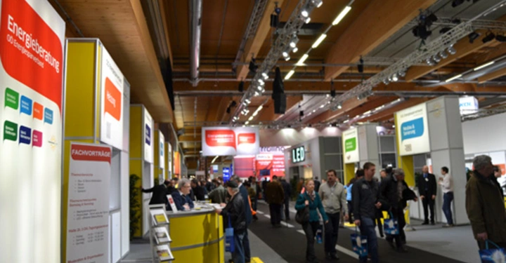 Galerie-Bild 33: Mehrere Stände befinden sich in einer gut beleuchteten Messehalle Menschen gehen durch die Gänge Schilder über den Ständen zeigen verschiedene Unternehmenslogos und Begriffe wie Energieberatung und Fachvorträge Lichter und Dekor oben