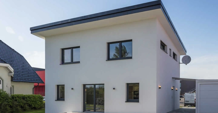 Modernes zweistöckiges Haus hat eine weiße Fassade große Fenster und ein Flachdach steht in einem gepflegten Vorstadtgarten andere Gebäude und ein blauer Himmel im Hintergrund.